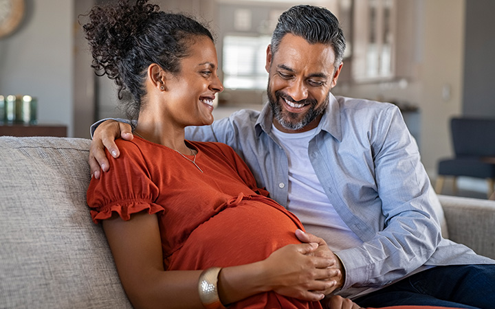 smiling man and woman on couch both holding her pregnant belly