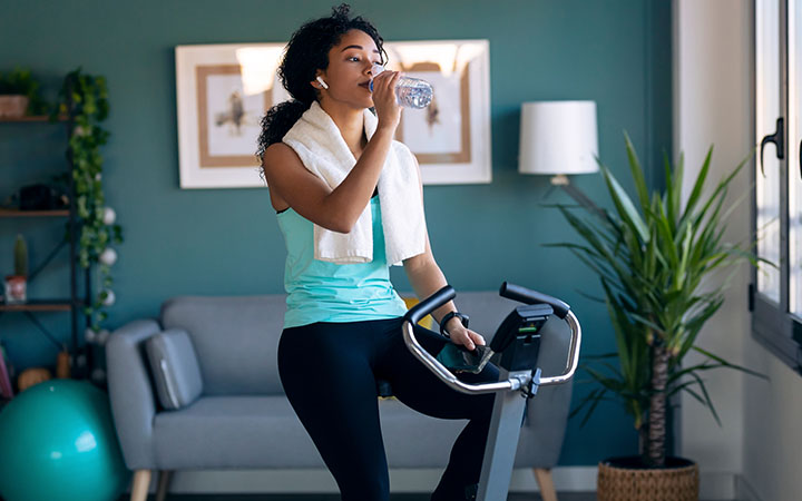 woman on stationary cycle drinking water from a bottle with towel around her neck