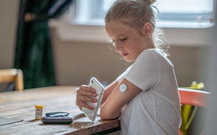 A young girl reads her blood sugar level by placing her cellphone next to her pump in her arm as she sits at a dining table
